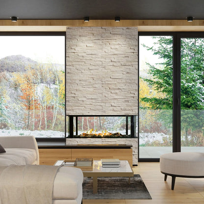A modern fireplace installation in a staged living room, featuring a tall rectangular stone veneer surround with a built-in linear firebox. The fireplace has a textured light beige stone face, a black glass front, and visible orange flames.