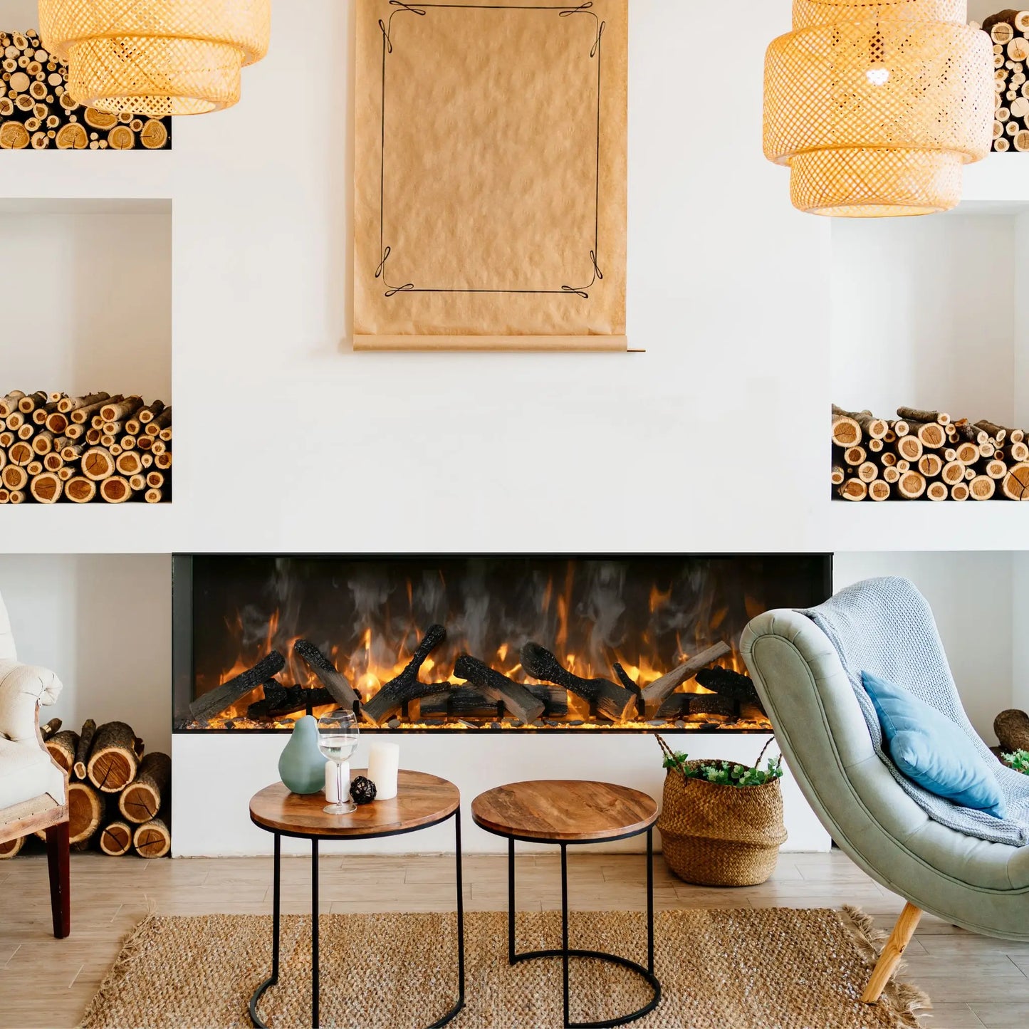 A 3-sided linear electric fireplace with a smoky orange flame effect set into a white wall between built-in niches stacked with firewood, under woven rattan pendant lights, with round nesting side tables and a light blue accent chair.