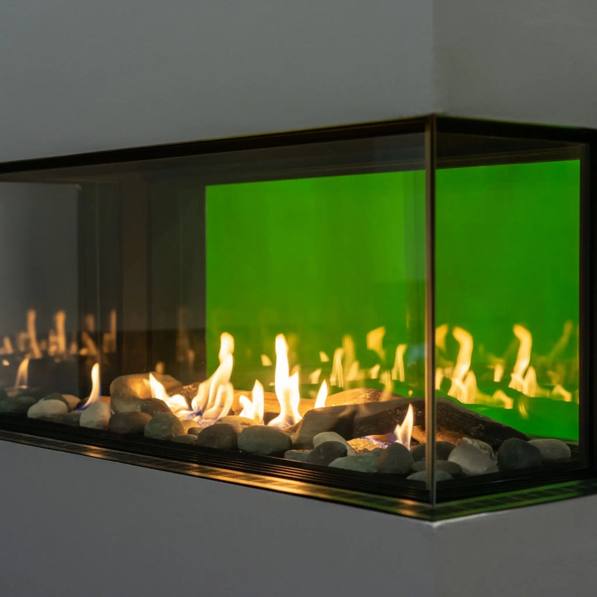 A modern linear fireplace insert with a black metal frame, clear glass front, glowing yellow-orange flames, and a vibrant green backlit interior, set against a neutral gray wall.
