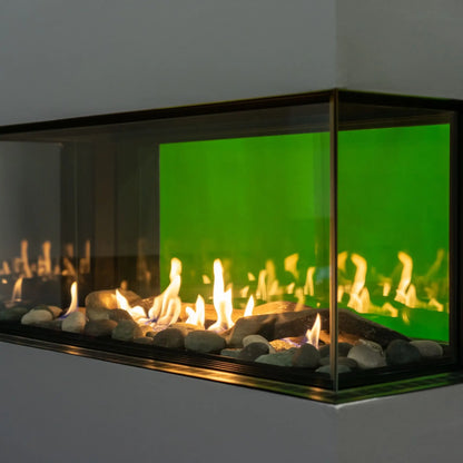 A modern linear fireplace insert with a black metal frame, clear glass front, glowing yellow-orange flames, and a vibrant green backlit interior, set against a neutral gray wall.