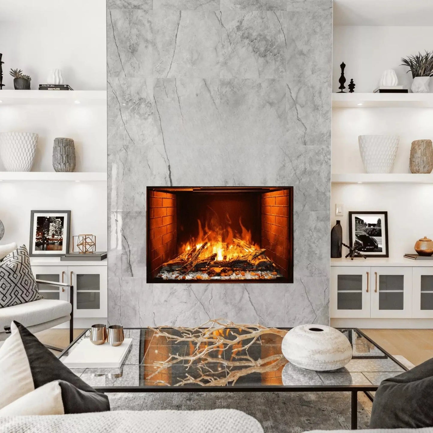 A built-in electric fireplace with orange flames set into a gray marble-tiled wall, flanked by white built-in cabinets with glass doors, above a glass coffee table styled with a branch centerpiece and a round ceramic vase.
