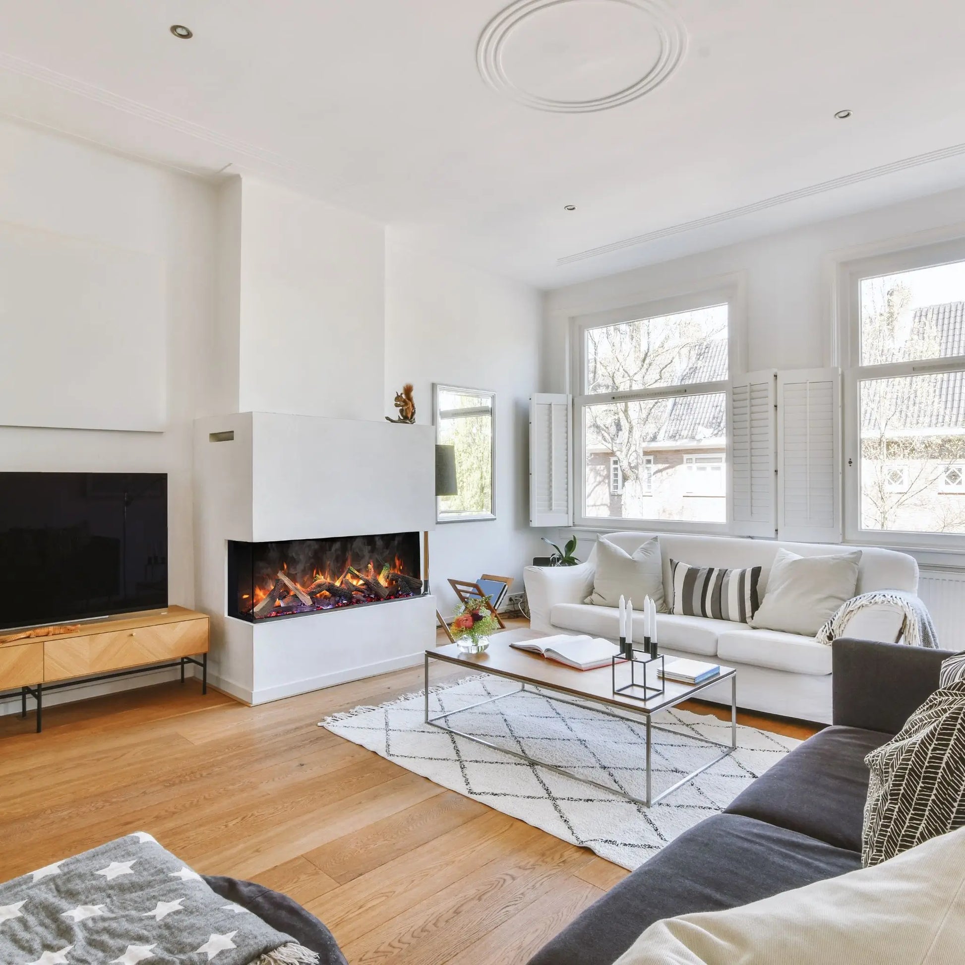A 3-sided linear electric fireplace with a glowing log set built into a white stone accent wall beneath a mounted TV, in a bright living room with tall windows, light wood floors, cream sofas, and a taxidermy squirrel figurine on the mantel.