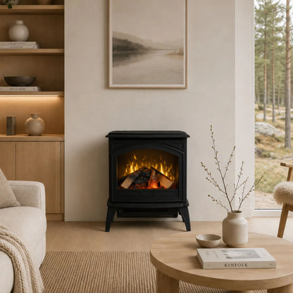 A black wood-burning fireplace with glowing orange flames and logs, set in a cozy, modern living room with a light wood coffee table, beige sofa, and a window showing a forest view.