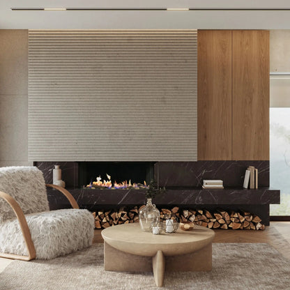 A modern living room interior featuring a large textured beige wall panel, a dark marble fireplace with visible wood logs, a round light wood coffee table, and a fluffy white armchair.