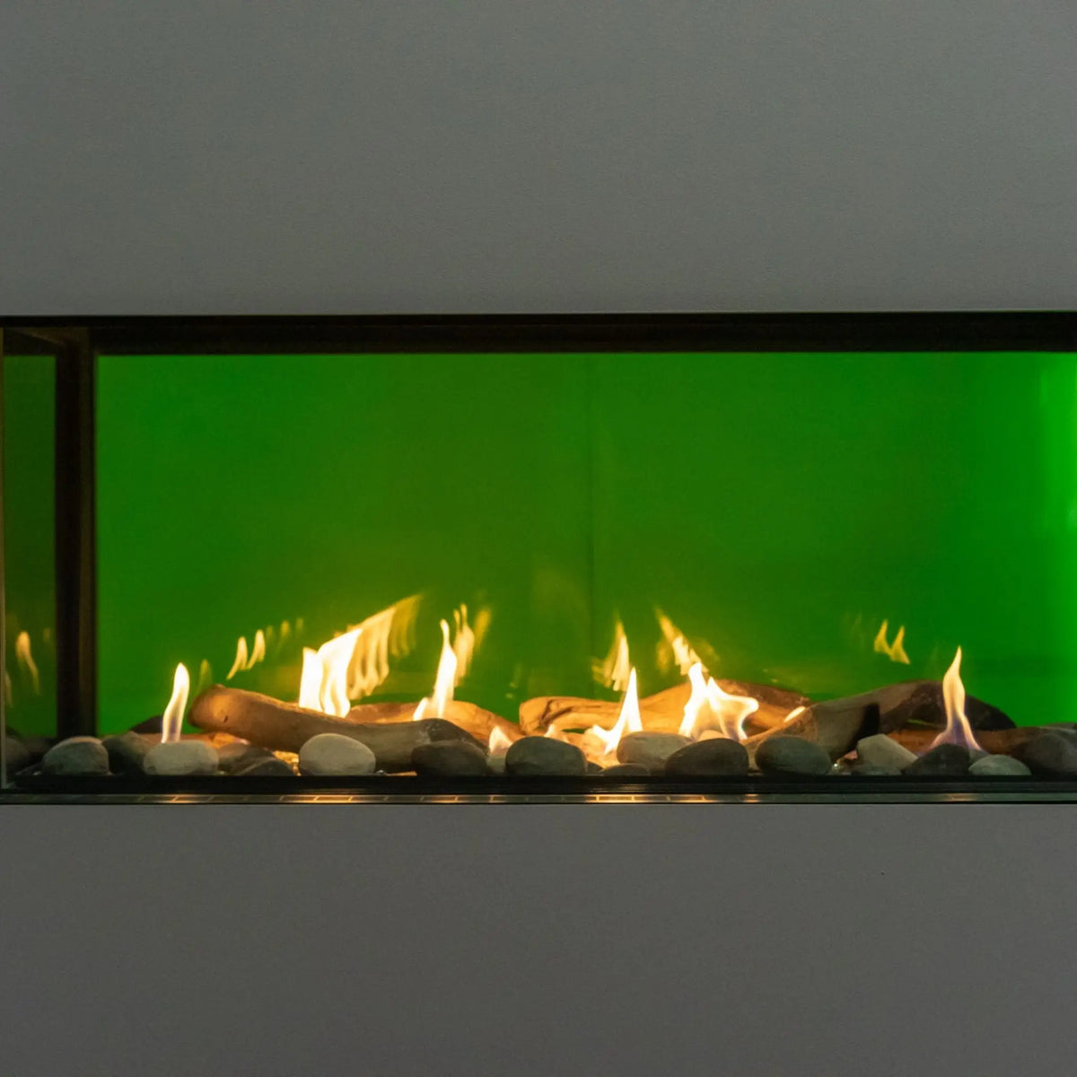 A modern linear fireplace insert with a vibrant green backlit glass panel, featuring glowing yellow and orange flames over dark wood logs and smooth gray stones.