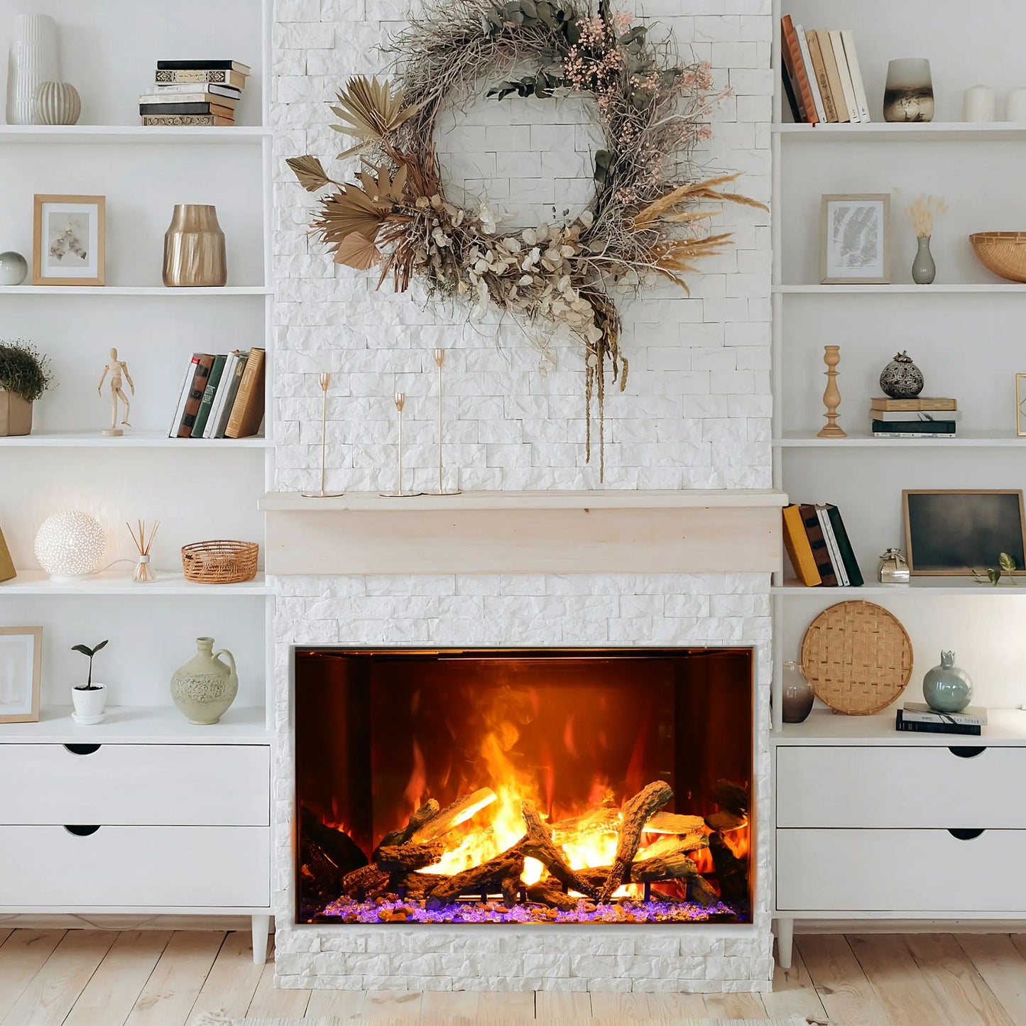 A built-in electric fireplace with orange flames and purple glass ember bed set into a whitewashed brick wall, styled with a large dried flower and pampas wreath above the mantel, flanked by white built-in bookshelves in a neutral boho living room.
