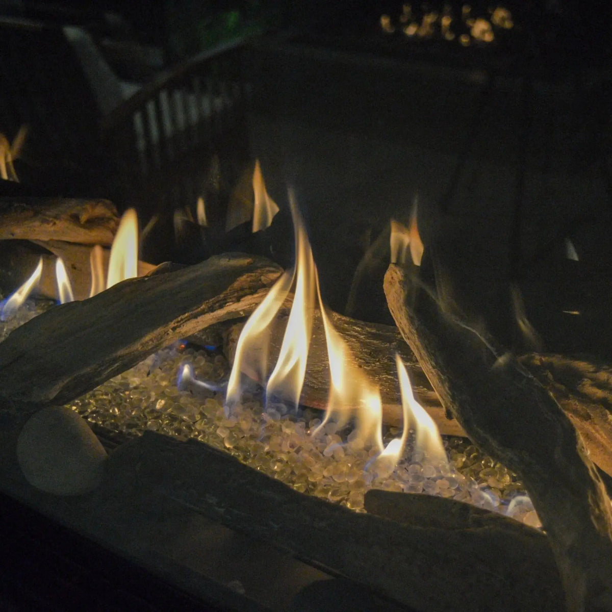 Gas fireplace with glowing yellow and orange flames rising over dark wood logs and clear glass stones in a dimly lit setting.
