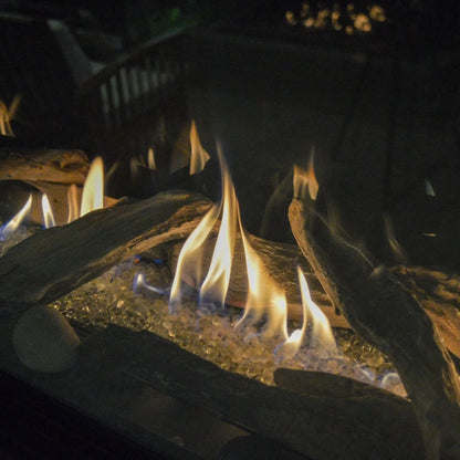 Gas fireplace with glowing yellow and orange flames rising over dark wood logs and clear glass stones in a dimly lit setting.
