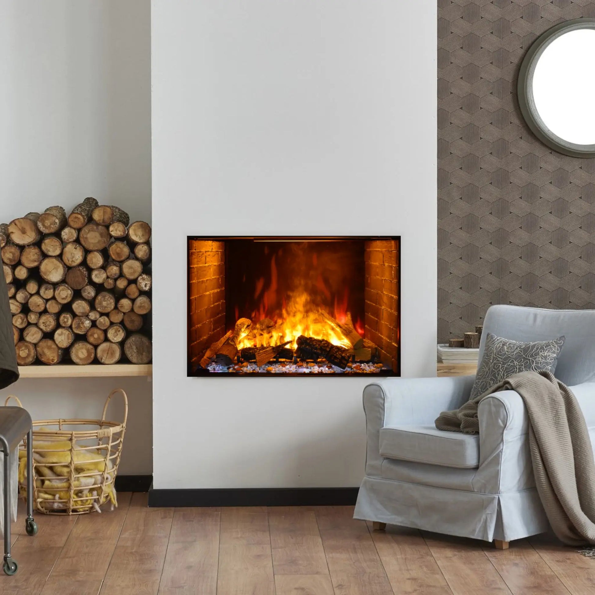 A built-in electric fireplace with an orange flame effect set into a tall white wall, flanked by a stacked firewood display on one side and a round mirror on the other, in a living room with gray patterned wallpaper and a light blue armchair.