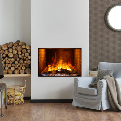 A built-in electric fireplace with an orange flame effect set into a tall white wall, flanked by a stacked firewood display on one side and a round mirror on the other, in a living room with gray patterned wallpaper and a light blue armchair.