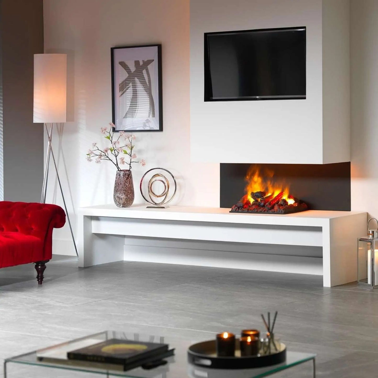 An Optimyst water vapor cassette installed in a simple black-framed opening on a white wall beneath a mounted TV, shown burning in a minimalist living room with a red velvet chaise and a floor lamp.