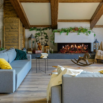 A 3-sided linear electric fireplace with orange flames set into a white wall niche framed by trailing greenery, in a rustic living room with exposed wood ceiling beams, gray sofas, yellow throw pillows, and a wooden rocking horse.