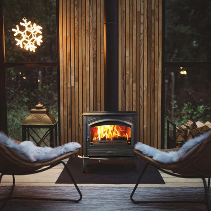 A black wood-burning fireplace with a glowing orange fire, set in a cozy room with wood-paneled walls, two chairs with white faux-fur cushions, and large windows showing a snowy light outside.