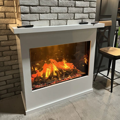 Angled view of the built-in electric fireplace installed in a white painted mantel surround with a floating shelf, two remote controls resting on top, next to a gray brick wall and wooden bar stools in a showroom setting.