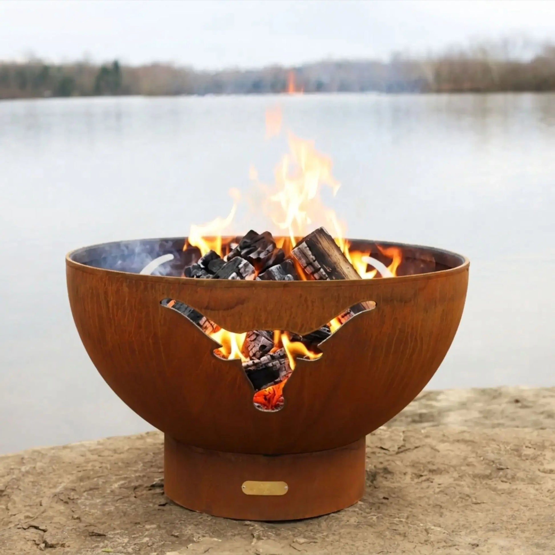 Rust-colored fire pit with flames burning on a stone surface by a body of water.