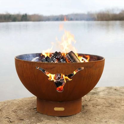 Rust-colored fire pit with flames burning on a stone surface by a body of water.