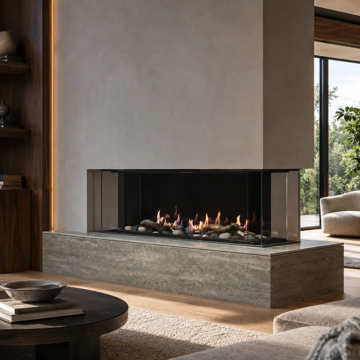 A modern linear fireplace with a concrete-look base and surround, glass front panels, and visible flames over dark logs and stones, set in a contemporary living room with wood flooring and a large window.