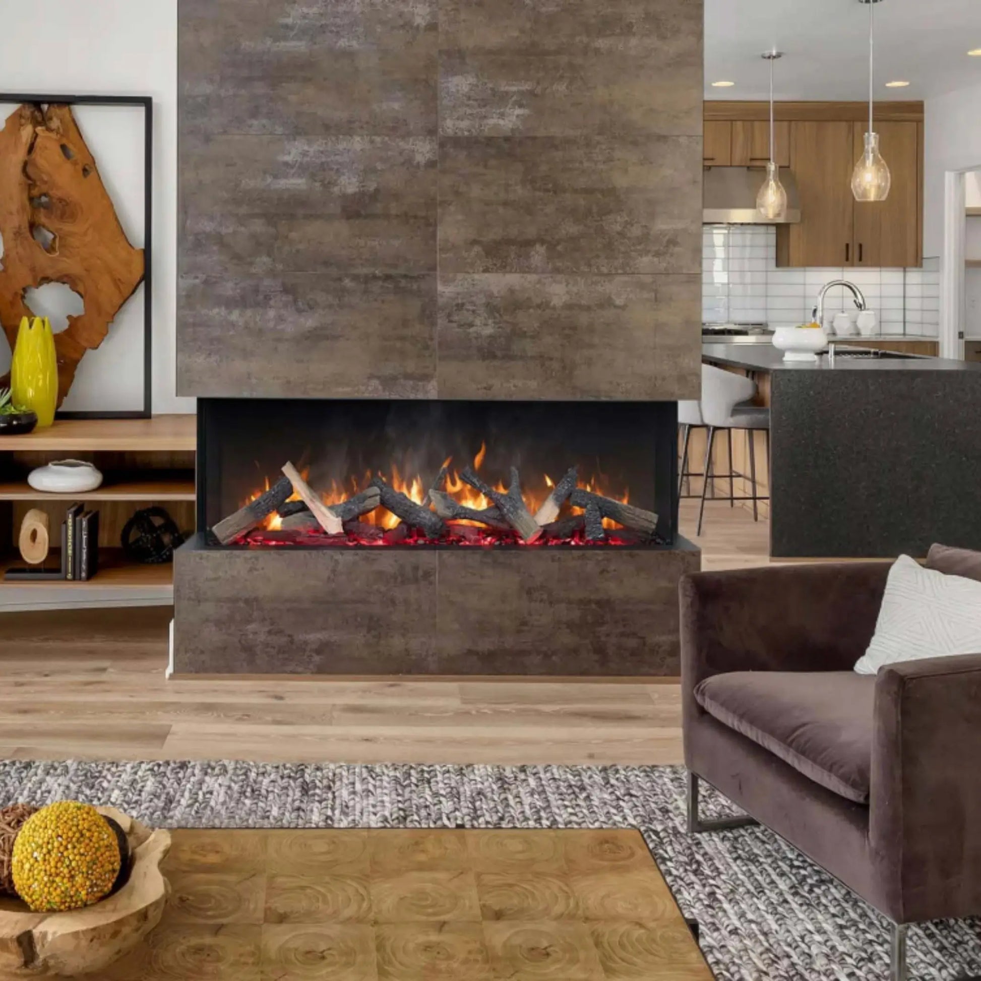 A 3-sided linear electric fireplace with a glowing log set built into a gray stone-tile wall, dividing an open-concept living room from a kitchen with pendant lighting, wood cabinetry, and a yellow vase on a nearby shelf.